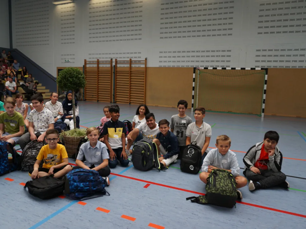 Kinder sitzen mit ihren Ranzen in der Sporthalle.
